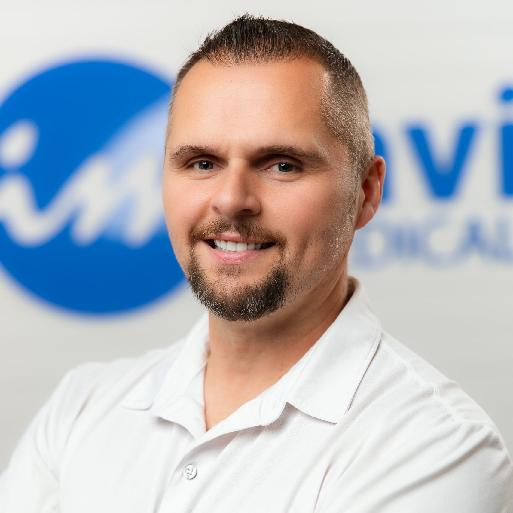 A man with short hair and a trimmed beard, wearing a white collared shirt, smiles confidently in front of a blurred blue and white logo background, reflecting his recent weight loss journey.