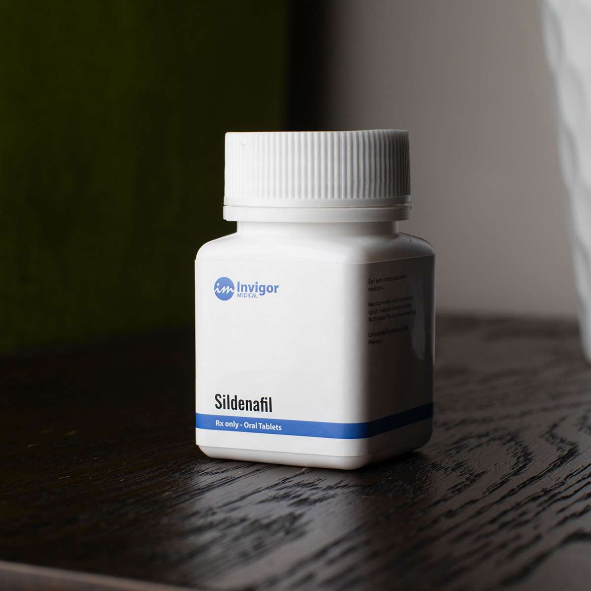 A white prescription bottle labeled Invigor Sildenafil, Rx only - Oral Tablets sits on a dark wooden surface with a green and white background—perfect for those looking to buy Enclomiphene online.